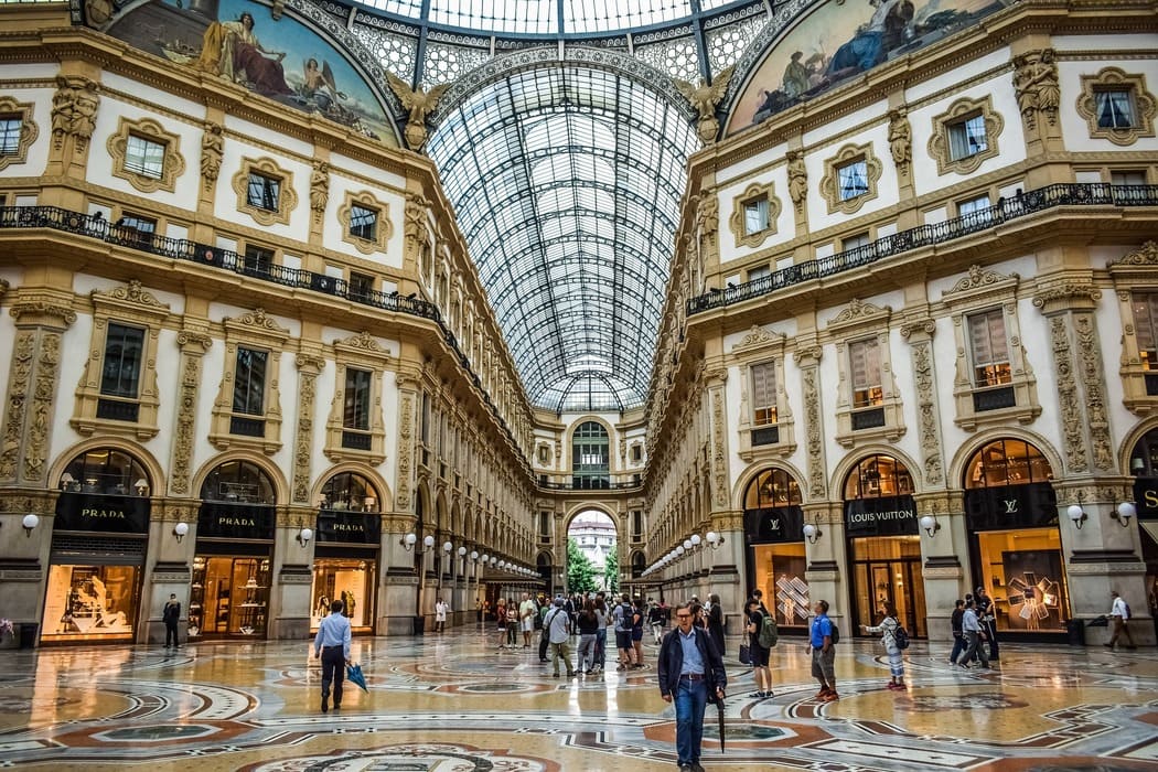 Atravessamento urbano na Galleria Vittorio Emanuele II, lugar bem iliminado com espaco amplo ladeado por vitrines de lojas.